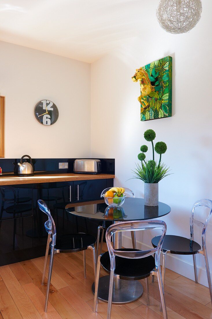 Small, round dining table and plexiglass chairs in modern kitchen with black kitchen counter and pale wooden floor