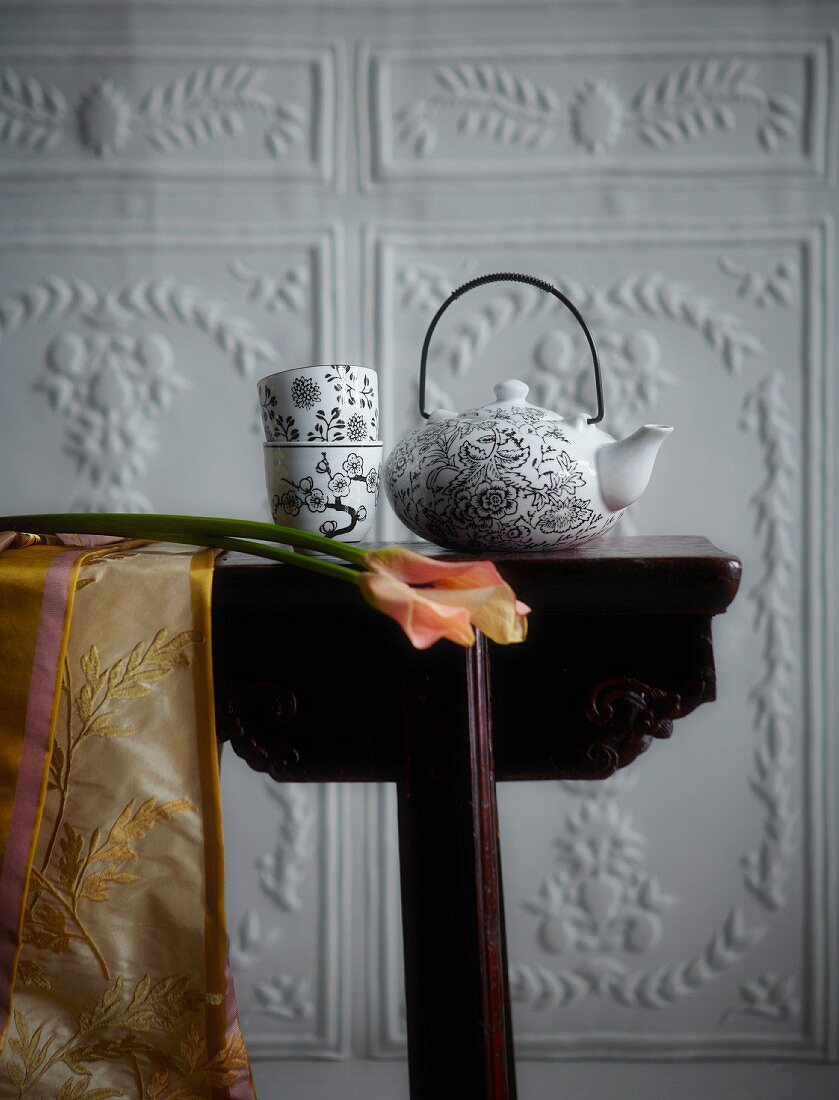 Dunkler Holztisch mit weiss-schwarz gemusterter Teekanne und Teebechern aus Porzellan vor weisser Wand mit floralen Reliefs