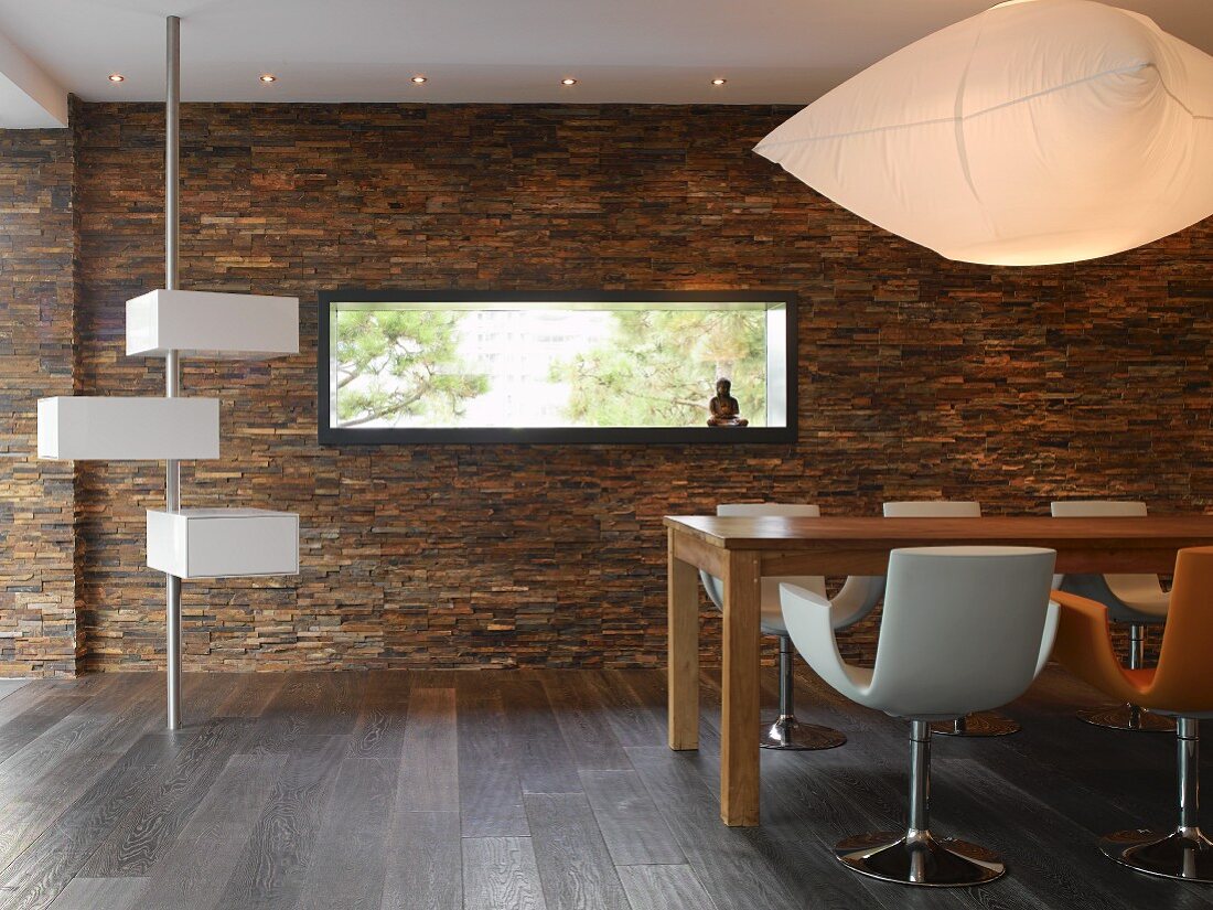 Designer Wohnraum mit weißen Aufbewahrungskästen an Metallstange daneben massiver Holztisch mit Schalenstühlen vor gefliester Wand mit Naturstein und rechteckiger Festverglasung