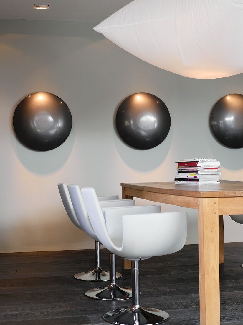 White designer chairs at massive wooden table on dark parquet floor with metal half-spheres under spotlights on white wall