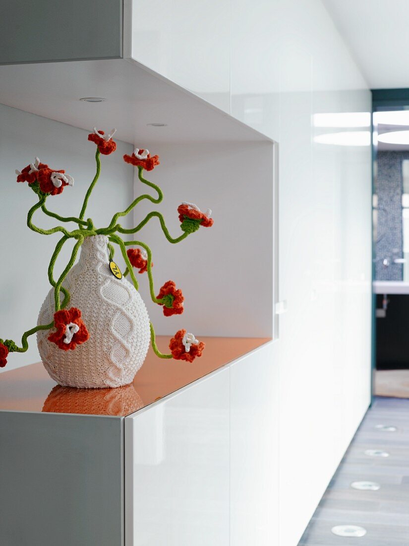 White, glossy, fitted cupboards with cut-out and orange stained glass shelf holding vase with crocheted cover and orange, crocheted flowers on green, wiggly stems