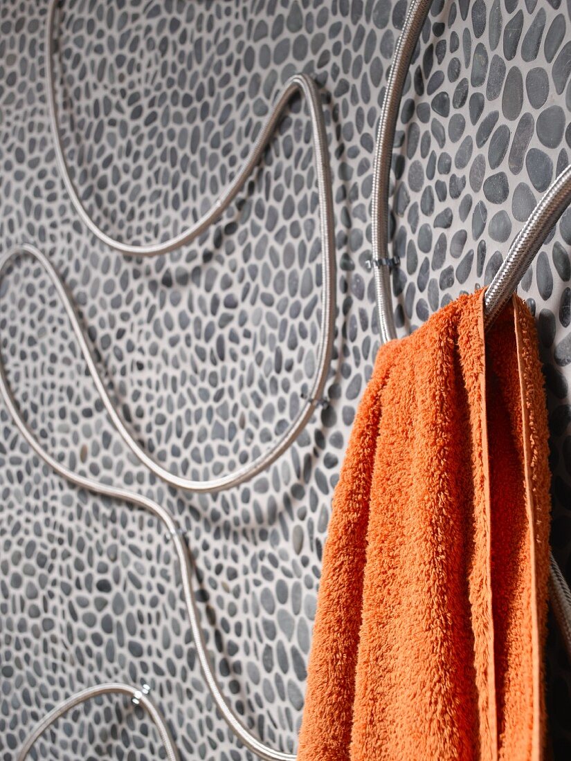 Curved shower hose mounted on pebble mosaic tiled wall and used as towel rail for orange towel