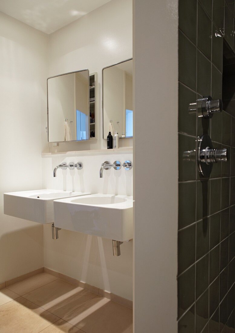 Designer bathroom with two white sinks, wall-mounted taps and mirrored wall cabinets; tiled wall with shower fittings in foreground