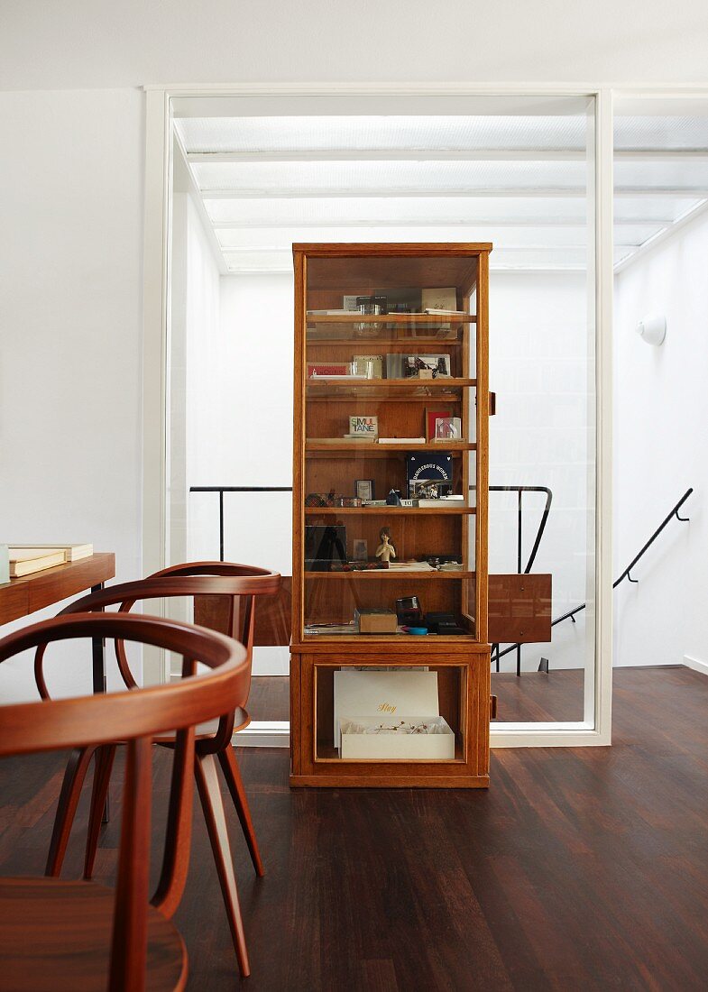 Holzvitrinenschrank steht vor Glaselement zum Treppenabgang auf dunklem Holzparkett, im Vordergrund Tisch mit eleganten dunklen Holzstühlen