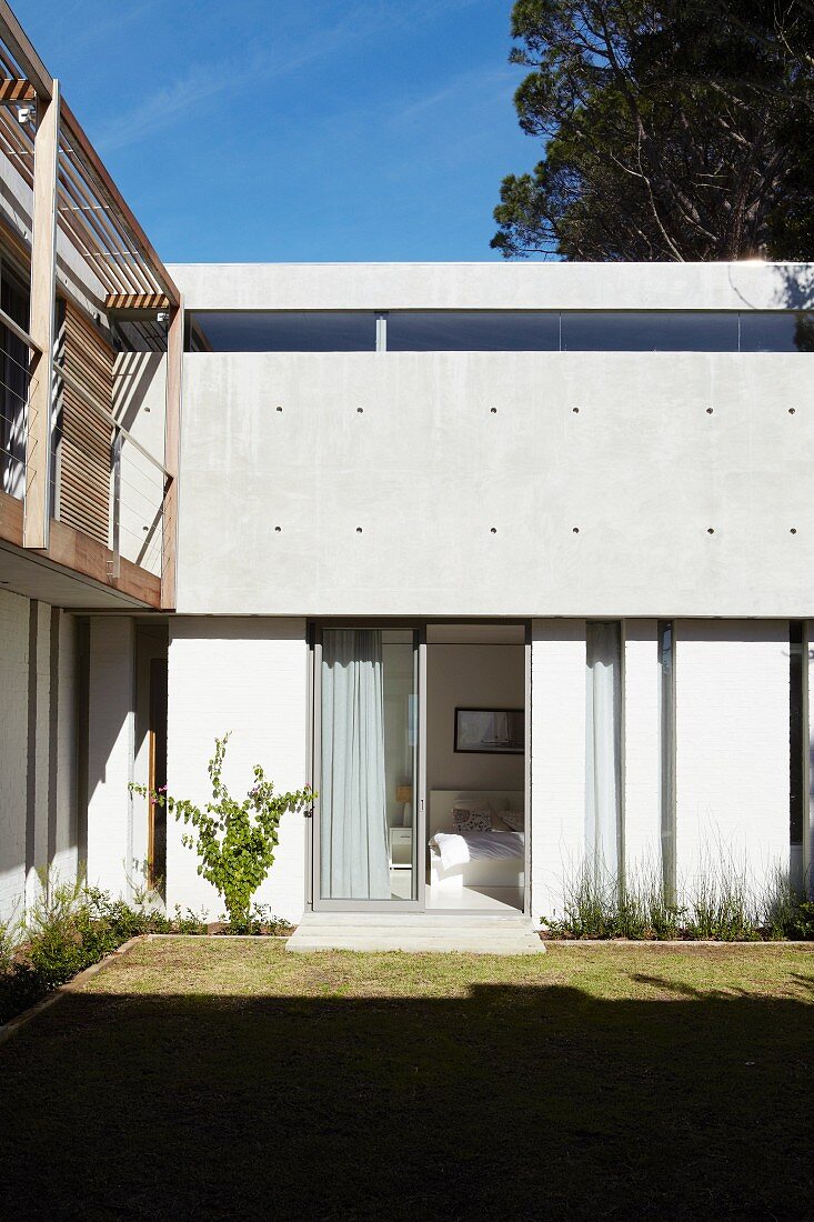Garden facade of modern residential house in mixture of materials with exposed concrete, white-painted wall and natural wood; view in through open bedroom window