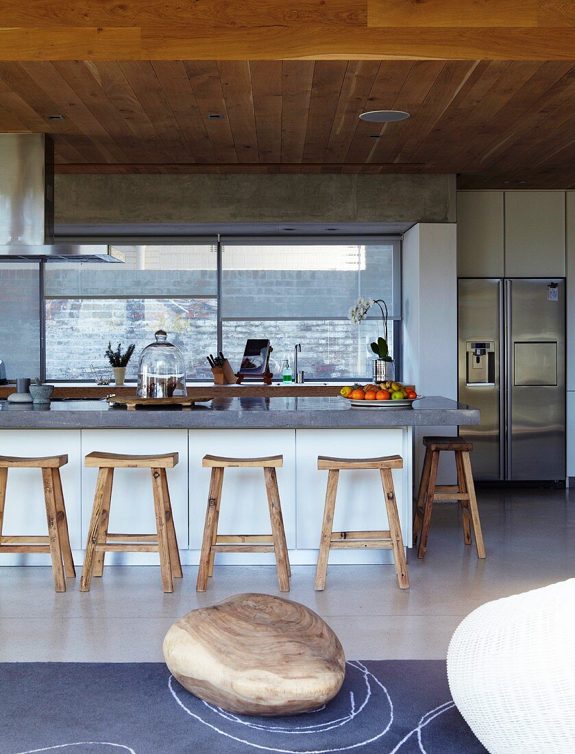 Puristische Küche mit rustikalen Holzhockern an der Frühstückstheke; Kunstobjekt aus einem Holzblock auf dem Schieferboden