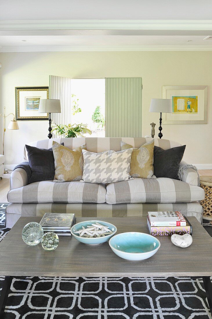 Comfortable sofa with grey and white striped upholstery and many scatter cushions behind coffee table