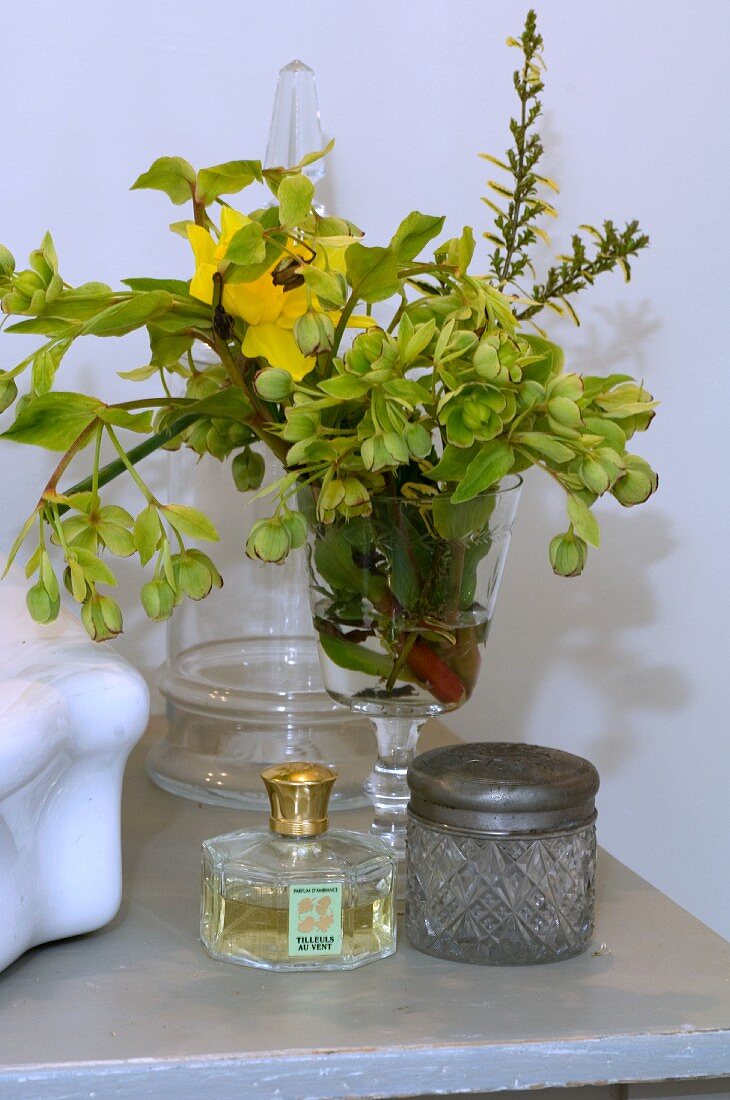 Blumensträusschen in einem Weinglas; davor ein Parfumflakon und ein Kristalldöschen mit Metalldeckel