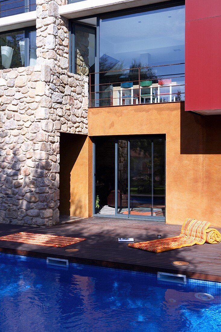 Modernes Haus mit Glasfronten und Natursteinmauern; davor ein azurblauer Swimmingpool mit Holzterrasse