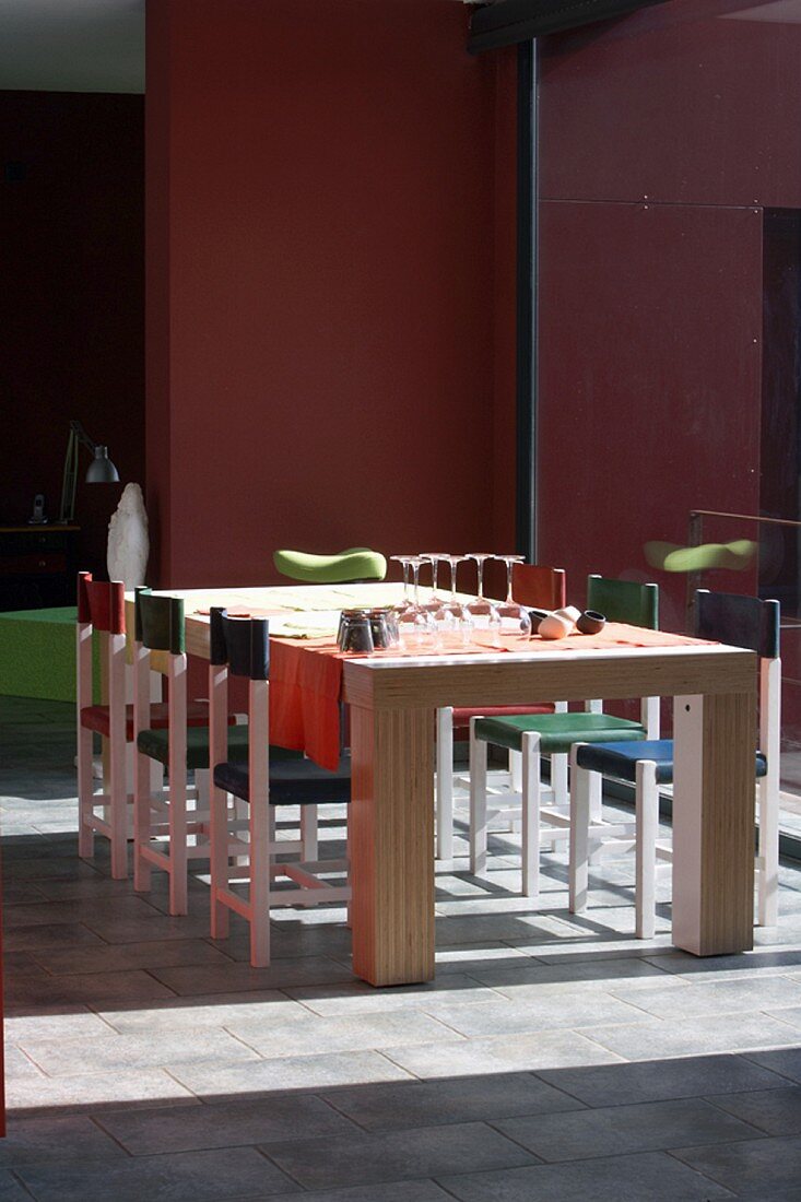 Chairs of various colours at solid wooden dining table next to floor-to-ceiling window