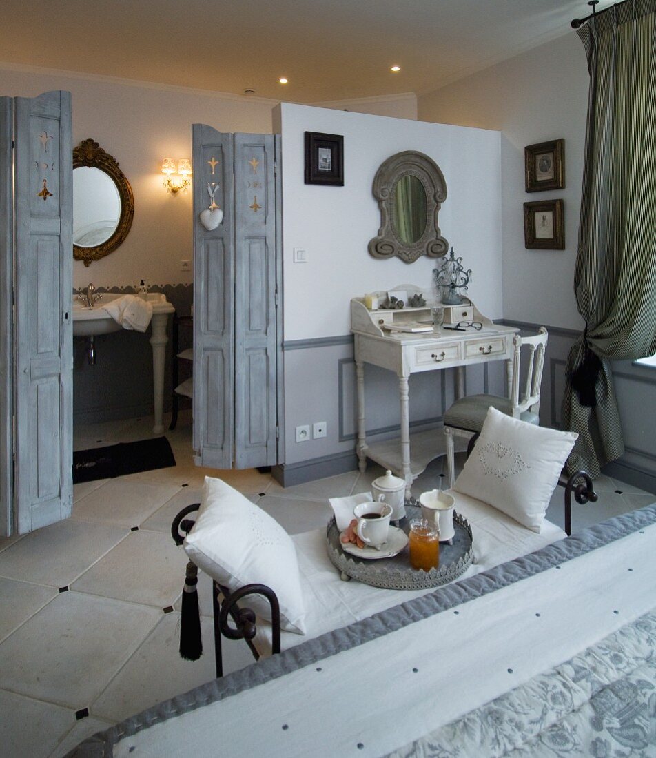 Blick vom Doppelbett mit kunstvoller Sitzbank und Frühstückstablett in Badezimmer mit grauer Falttüre