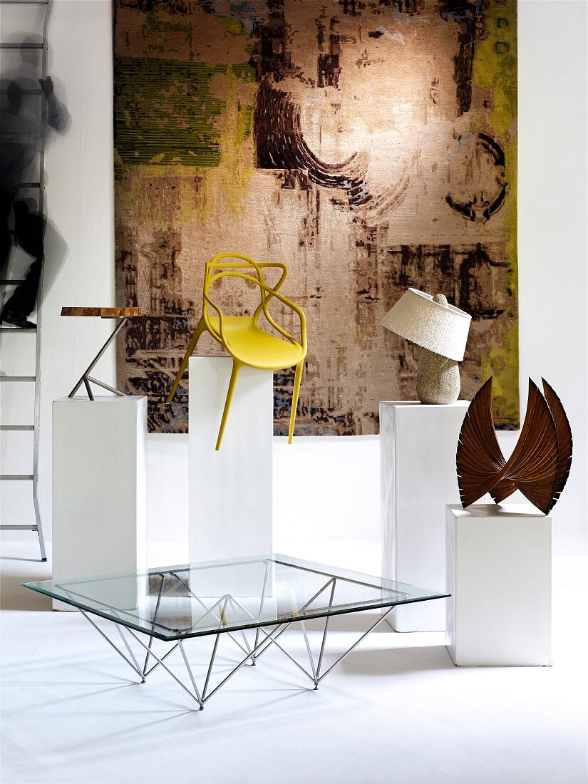 Stühle, Keramikgefäss und Skulptur auf Podest-Säulen und ein Glastisch vor Wandteppich