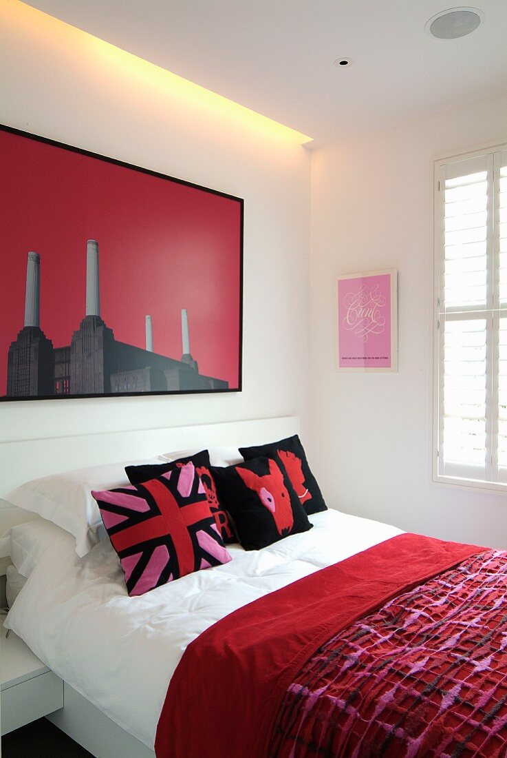 Rote gemusterte Tagesdecke auf Bett und modernes Bild an weisser Wand mit indirekter Beleuchtung
