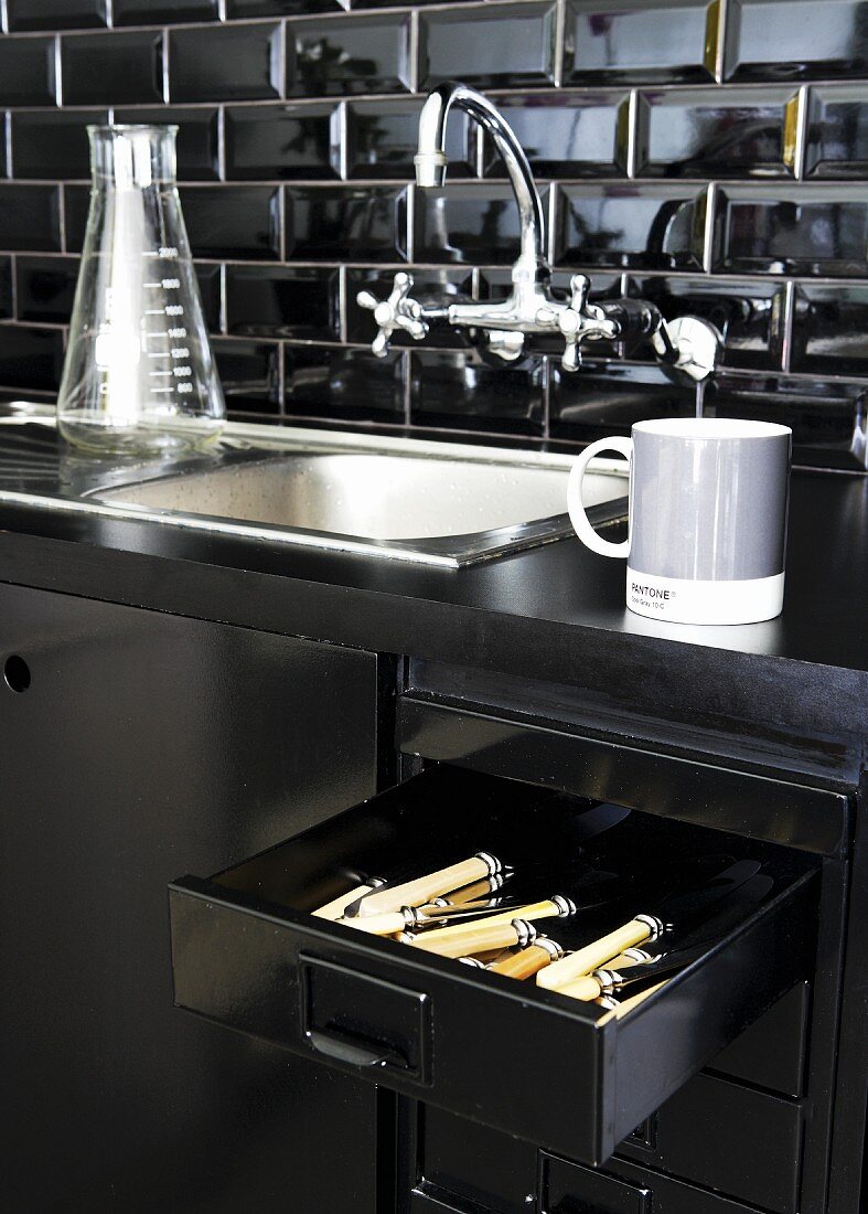 Detail of black kitchen counter with vintage-look sink and open cutlery drawer in base unit