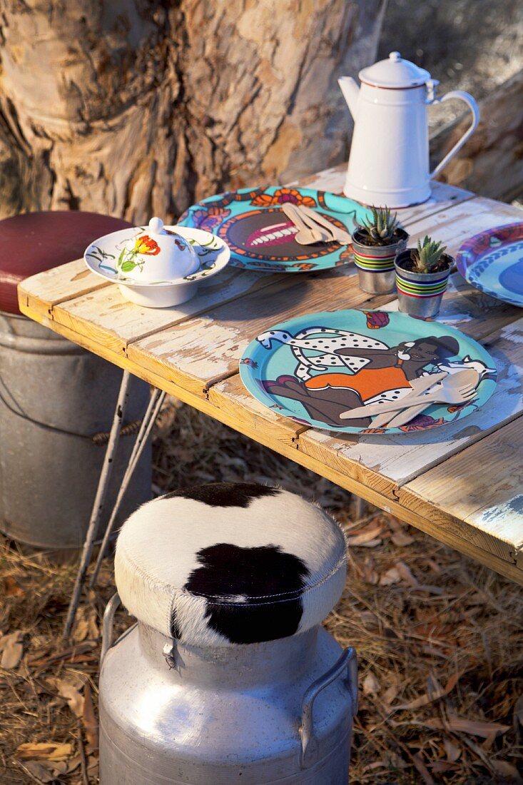 Gedeckter Tisch mit bemalten Tellern auf rustikaler Holzplatte und fellbezogene Vintage Milchkannen im Freien
