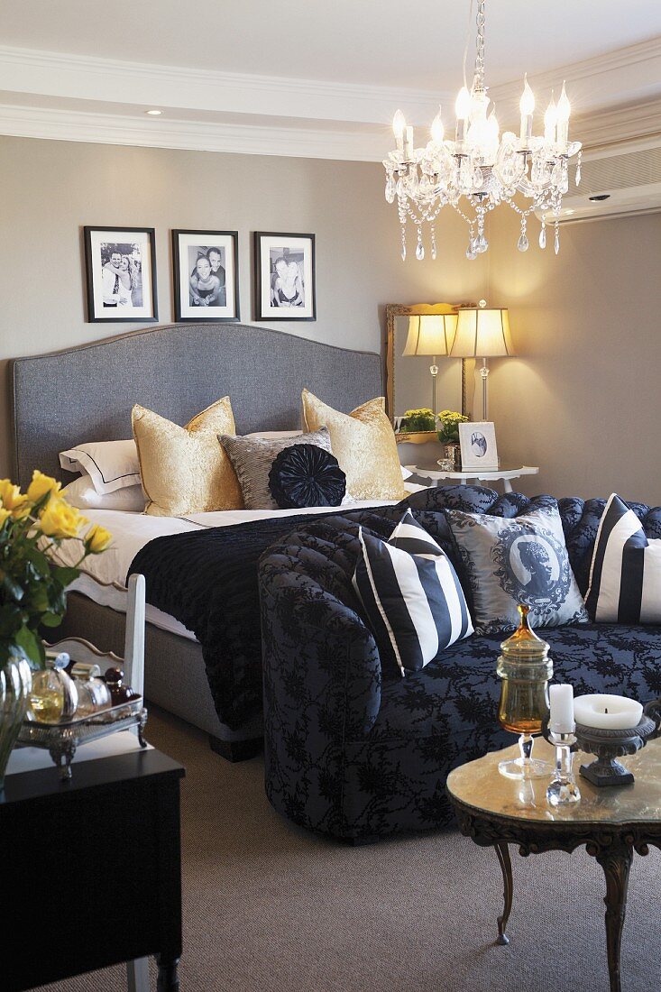 Traditionelles Schlafzimmer mit grauem Doppelbett und davorstehendem, schwarzen Polstersofa; weiche Zierkissen sorgen für Gemütlichkeit