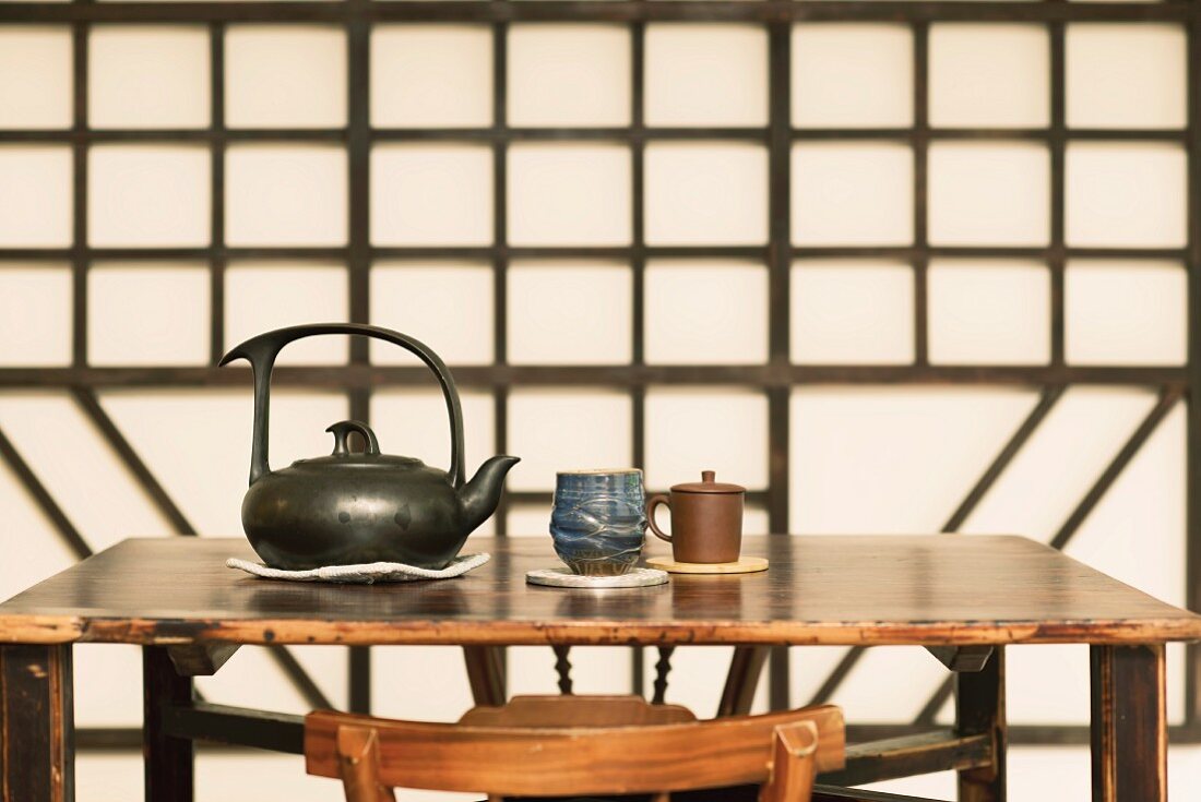 Ceramic teapot and teacups on Chinese-style wooden table: geometric wooden wall decoration in background