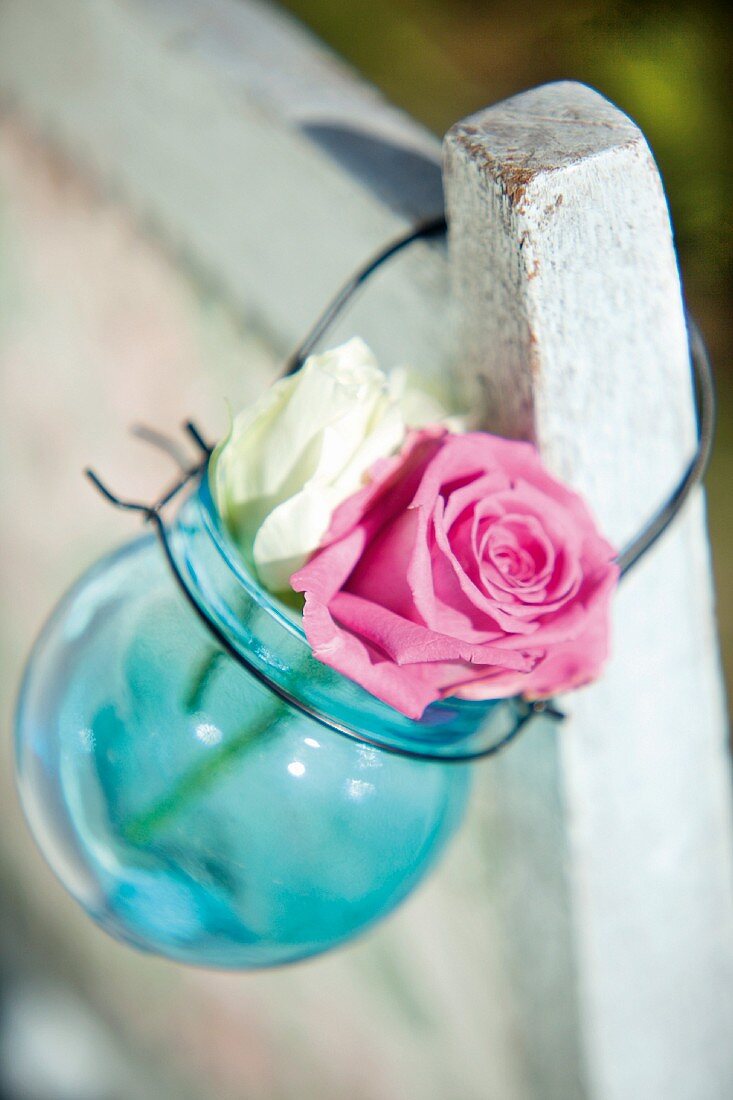 Zwei Rosenblüten in blauer Glaskugel mit Drahthenkel an Stuhllehne hängend