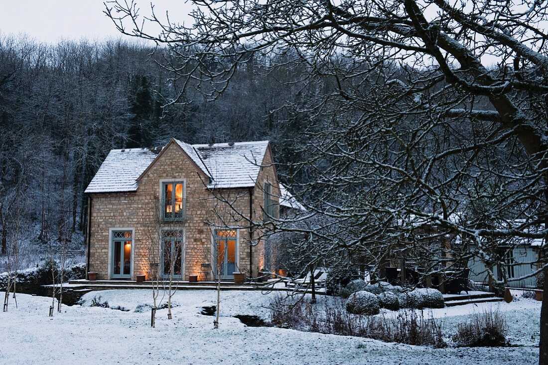 Blick über einen verschneiten Bachlauf auf das abendlich beleuchtete Verwalterhaus eines englischen Anwesens