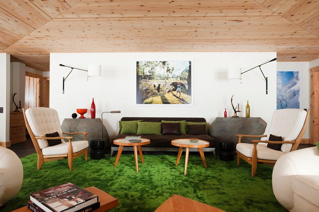 50s-style armchairs and side tables on green rug against wall with modern sconce lamps in interior with wooden ceiling