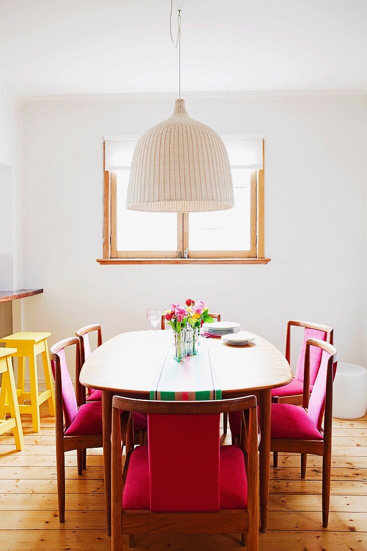 Dining table and wooden chairs with pink upholstery on stripped wooden floorboards in front of window in white wall