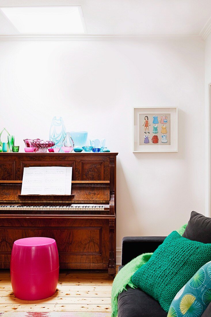 Collection of glass vessels on piano with pink barrel-shaped stool in corner of living room against white wall; black sofa in foreground