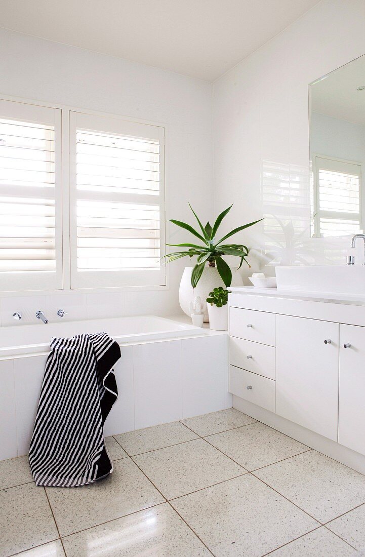 White washstand and bathtub in corner of bathroom below window with interior shutters of white slats