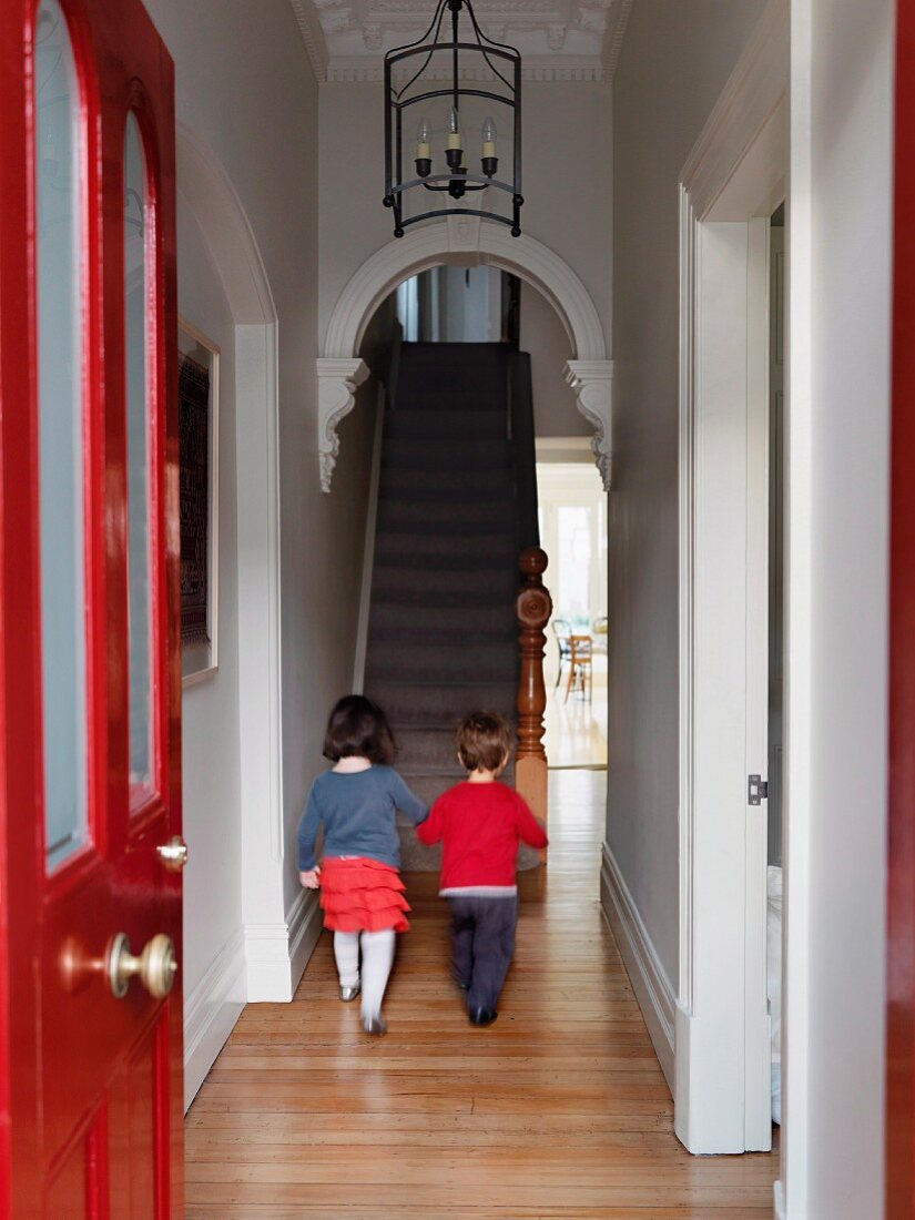 View through red-painted front door - two children walking along narrow hallway towards staircase