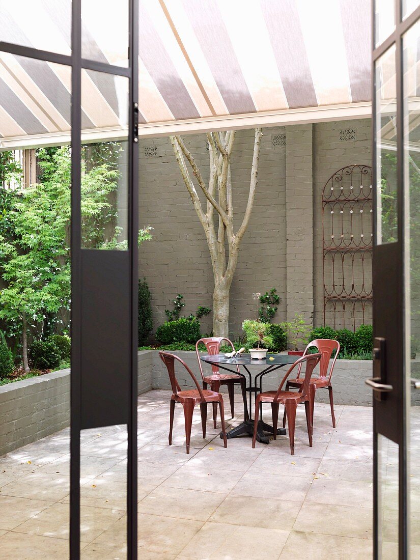 Blick durch offene Terrassentür auf farbige Metallstühle mit Retro-Flair an rundem Tisch im Innenhof