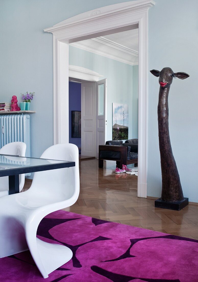 Zimmerflucht vom Essplatz ins Wohnzimmer; weiße 60er Jahre Stuhlklassiker auf knalligem Teppich und Giraffenskulptur im Hintergrund