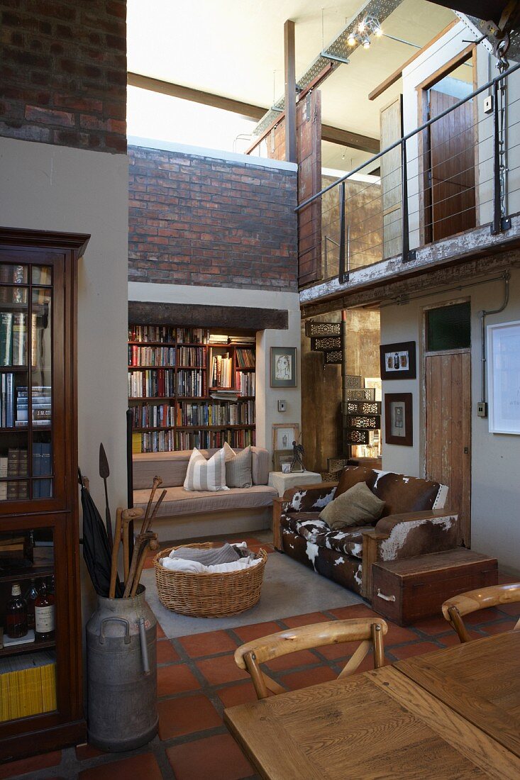 High-ceilinged interior with many reclaimed elements and gallery; sofa with animal-skin cover, old milk cans and library in background