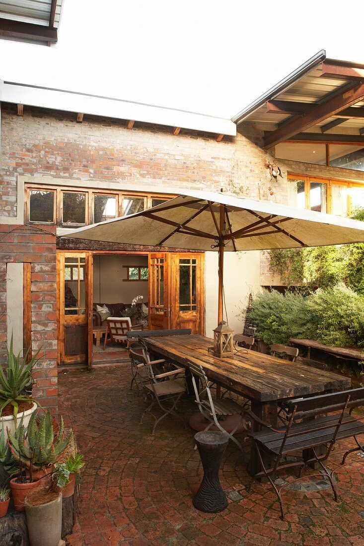 Seating area on terrace with market parasol in solid wooden table and old garden chairs; unconventional house with reclaimed elements in background