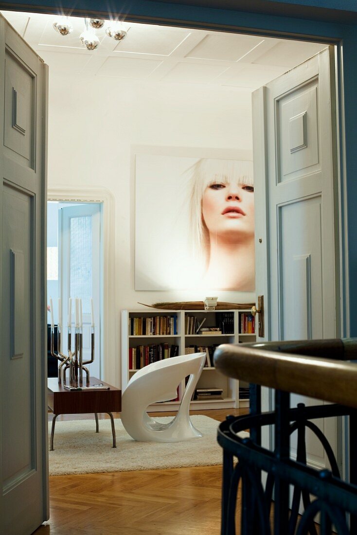 View through open double doors of white designer chair next to retro coffee table and large portrait of woman in classic setting