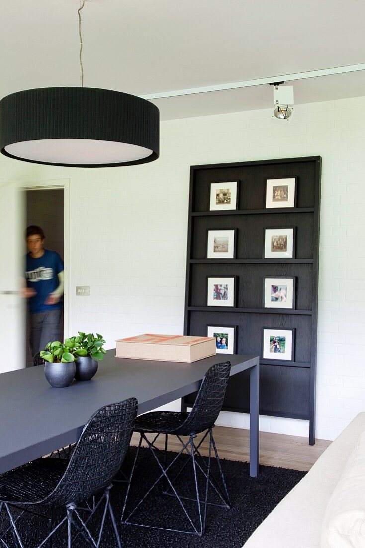 Anthrazitfarbener Esstisch mit geflochtenen Schalenstühlen; an der Wand ein schwarzes Paneel mit einer Fotosammlung