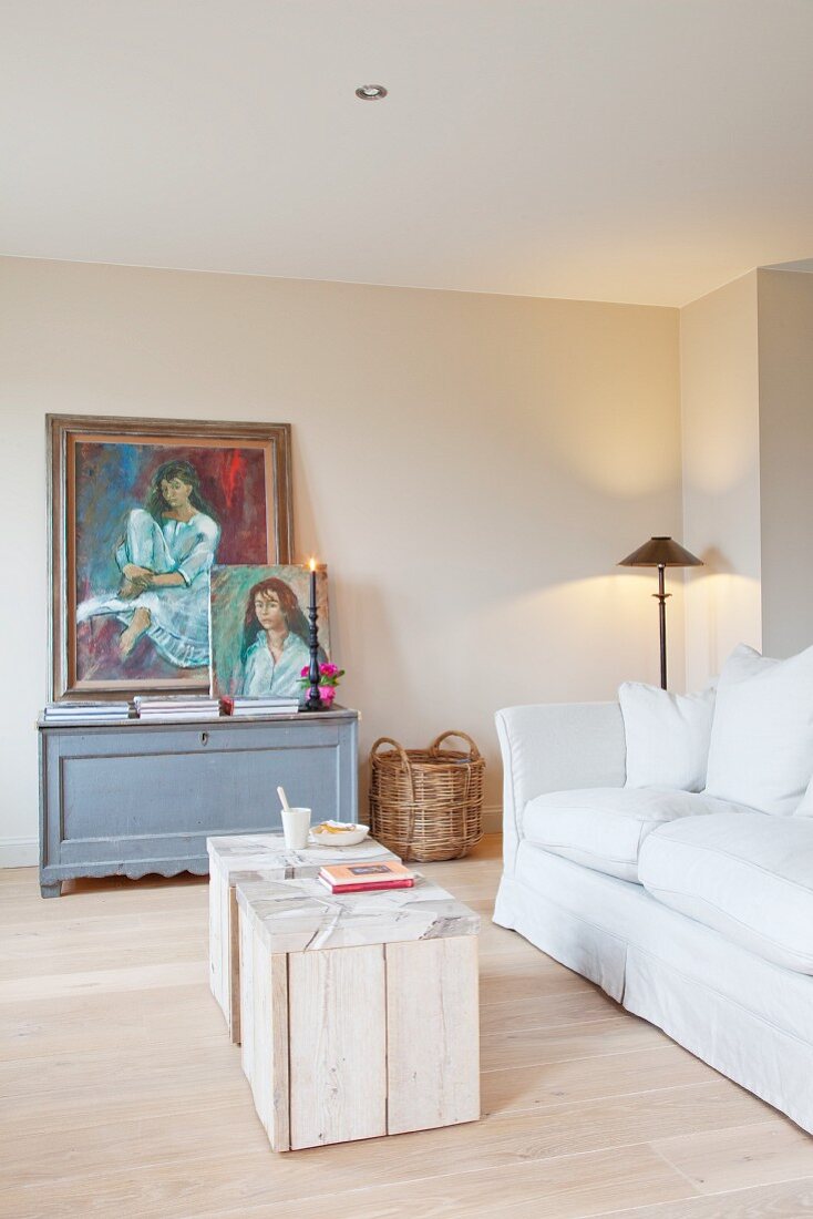 weiße Polstercouch mit zwei selbst gestalteten, rustikalen Couchtischen in hellem Wohnzimmer; im Hintergrund zwei Ölportraits auf einer grauen Vintage-Truhe