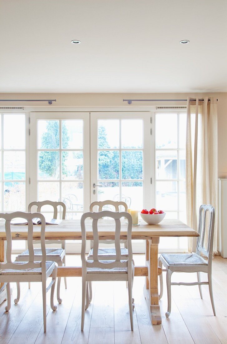 Muted dining room in clear natural shades with floor-to-ceiling windows, solid, pale wooden table and white vintage chairs