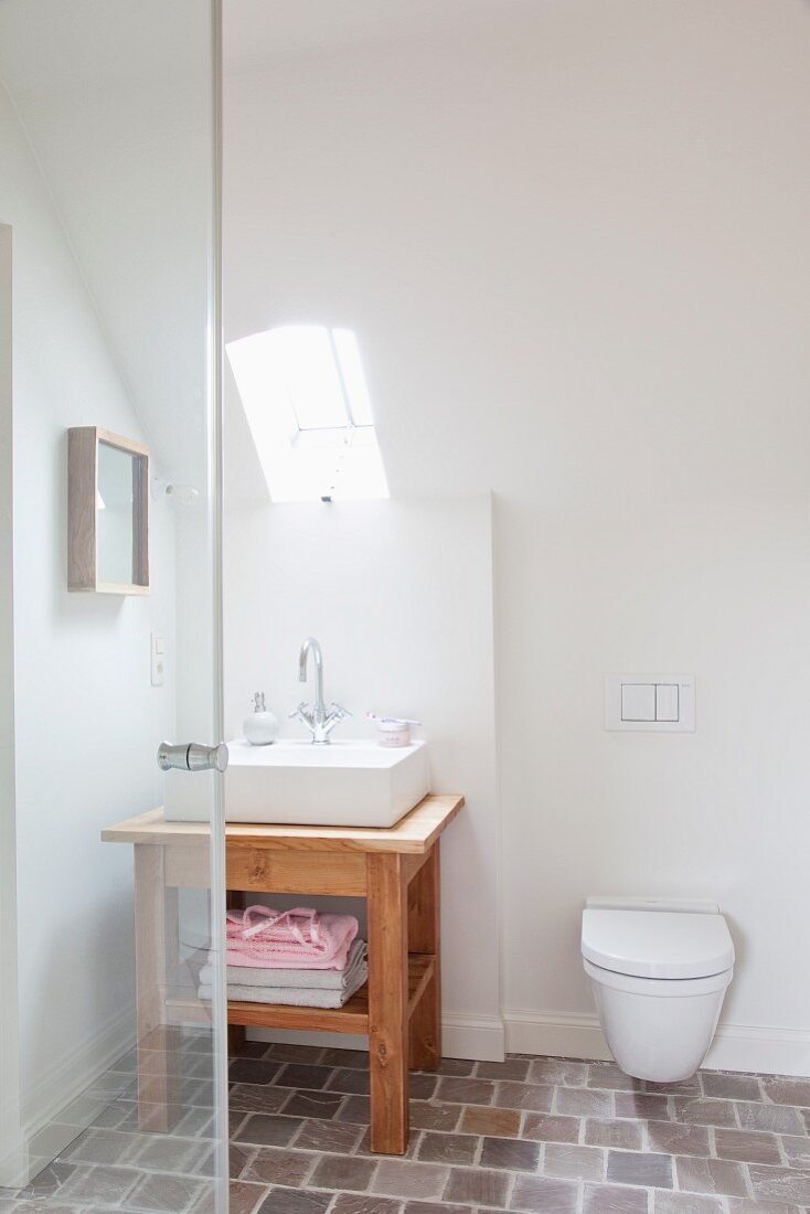 Wall-mounted toilet under sloping ceiling and small skylight above wooden washstand with rectangular basin