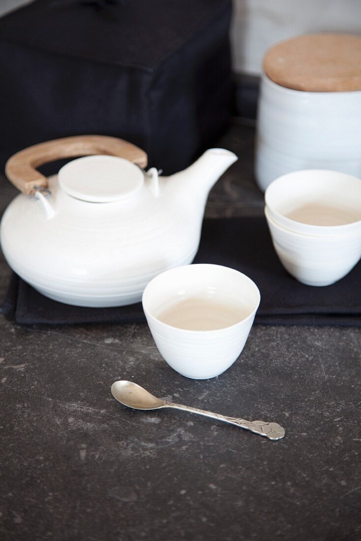 Weisses Teegeschirr auf grauem Naturstein, bestehend aus Teekanne mit Holzgriff, Teeschalen und Zuckerdose mit Holzdeckel