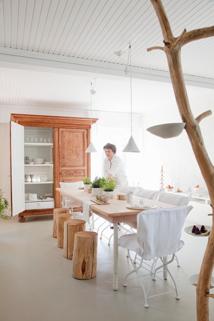 Woman setting dining table with tree stump stools and white garden chairs; wood and porcelain artwork in foreground