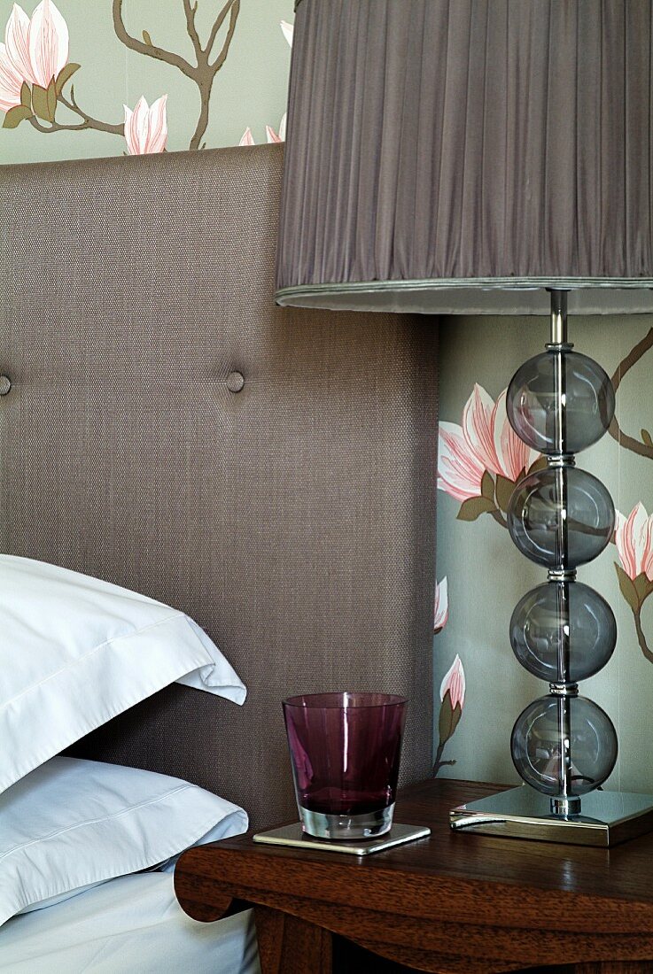 Coloured drinking glass next to table lamp with fabric shade on bedside table next to bed with upholstered headboard