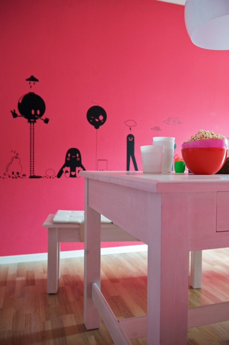 Schwarze Zeichnungen auf pinkfarbener Wand; davor ein weisser Holztisch mit Sitzbank