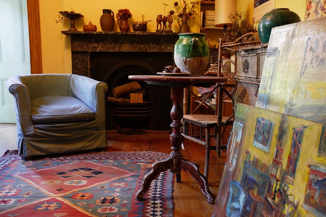 Corner of vintage-style living room - armchair with loose cover in front of open fireplace and wooden side table next to stack of paintings