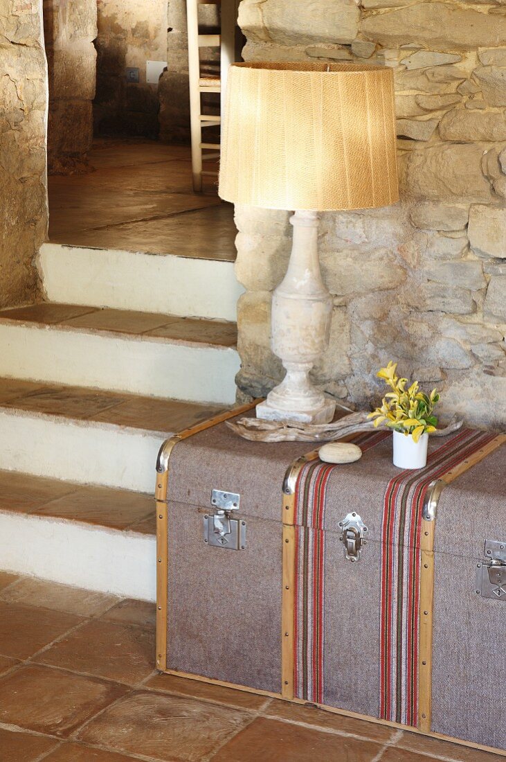 Traditionelle Tischlampe auf Reisekoffer vor Natursteinwand; Treppenstufen mit Terracottafliesen