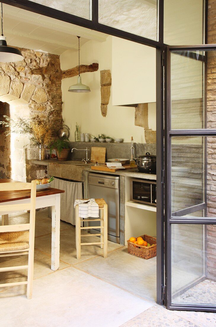 Blick durch geöffnete Glasfront auf einfache Küchenzeile und Essplatz mit mediterranen Geflechtstühlen