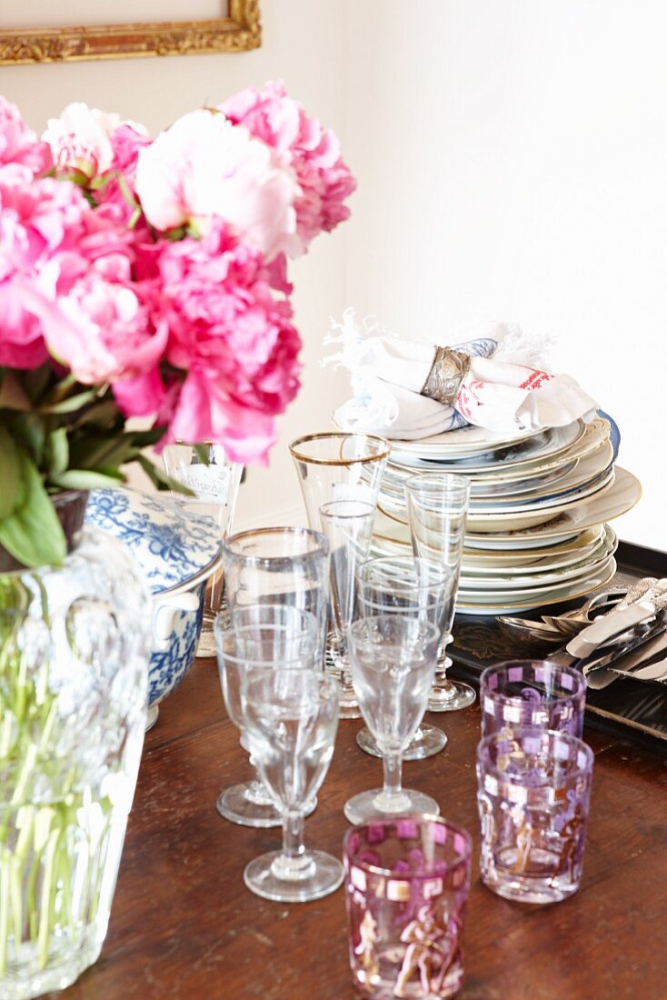 Stacked plates, various glasses and bouquet of peonies on wooden table