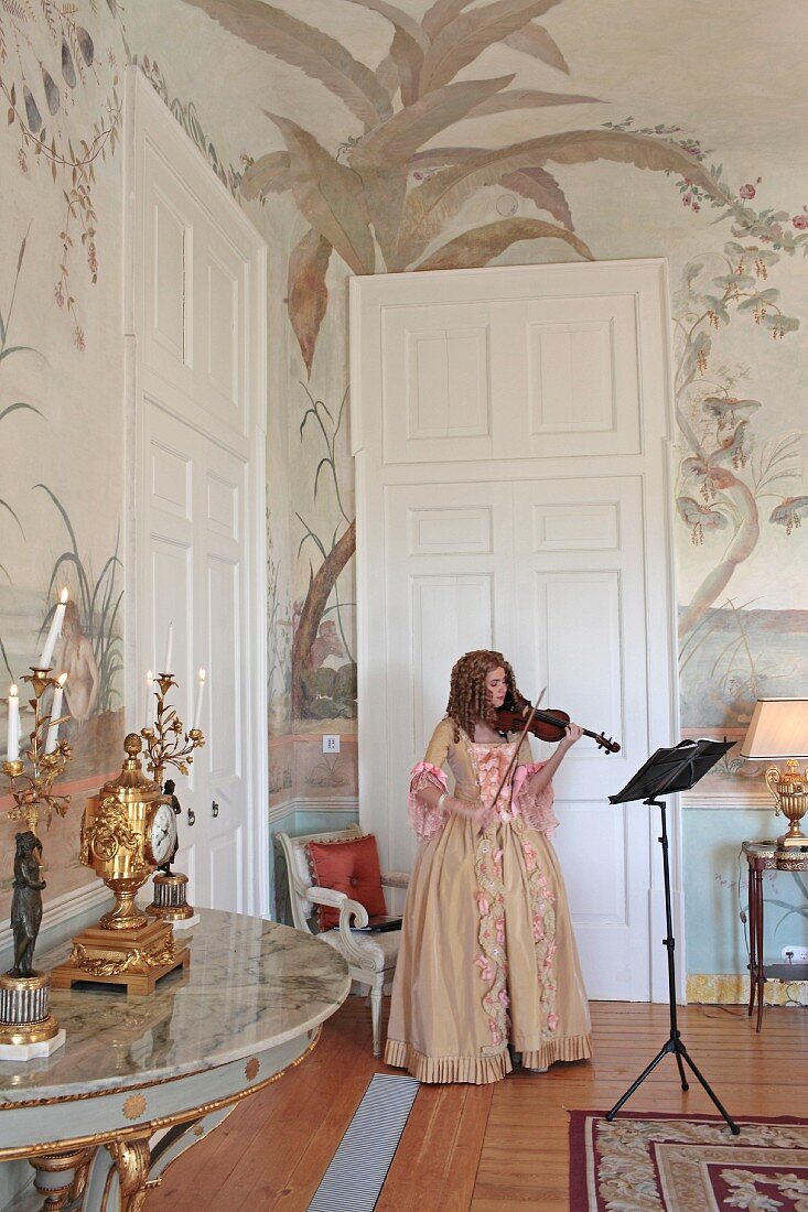 Frau mit antikem Kleid und Geige spielend in Salonecke mit bemalten Wänden