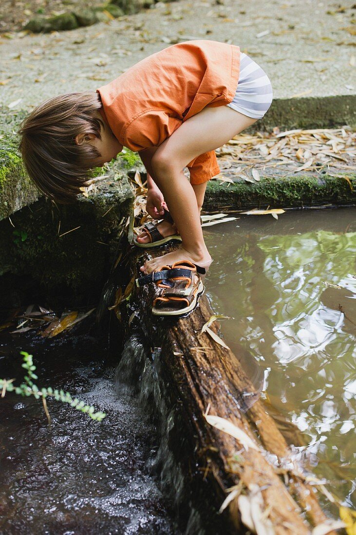 Boy putting on sandals on tree trunk across stream