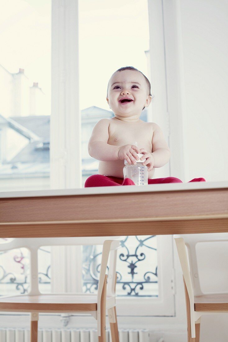Lachendes Baby sitzt mit seinem Fläschchen auf einem Tisch