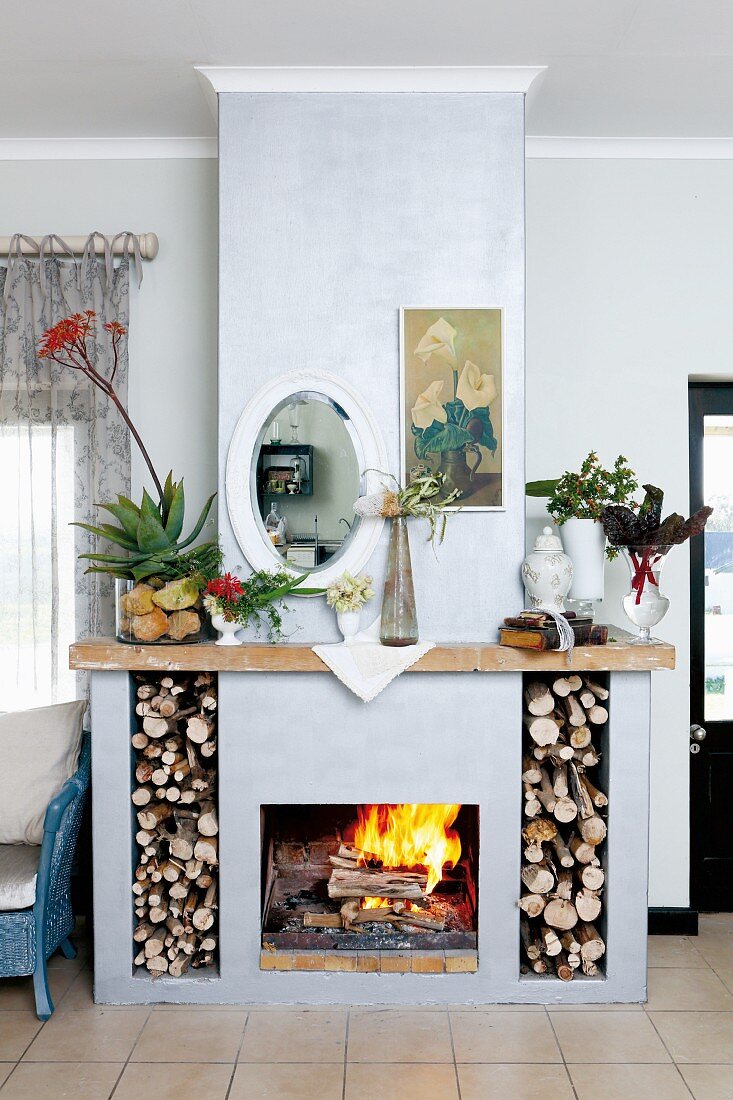 Mirror and floral picture hung above open fireplace with compartments for firewood and aloe and garden plants on mantlepiece