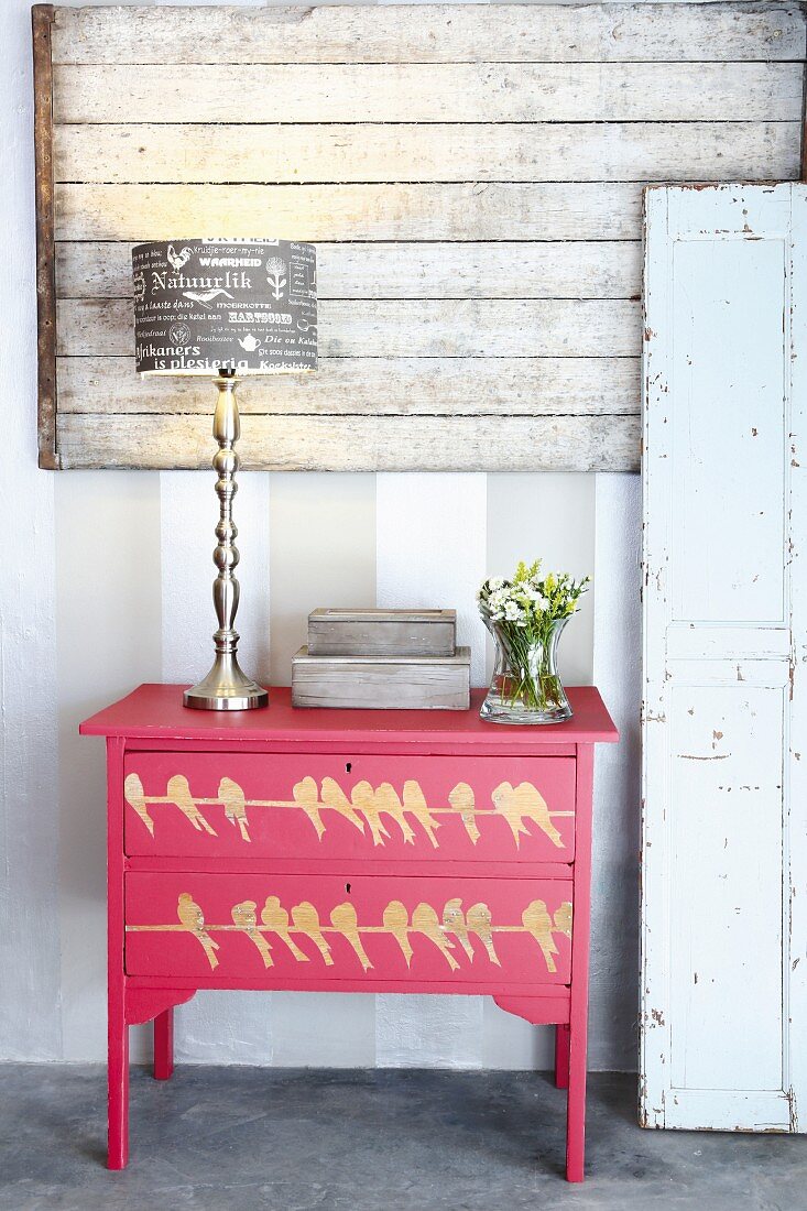 Lamp with printed paper shade, box and vase of flowers on chest of drawers painted red with bird motif